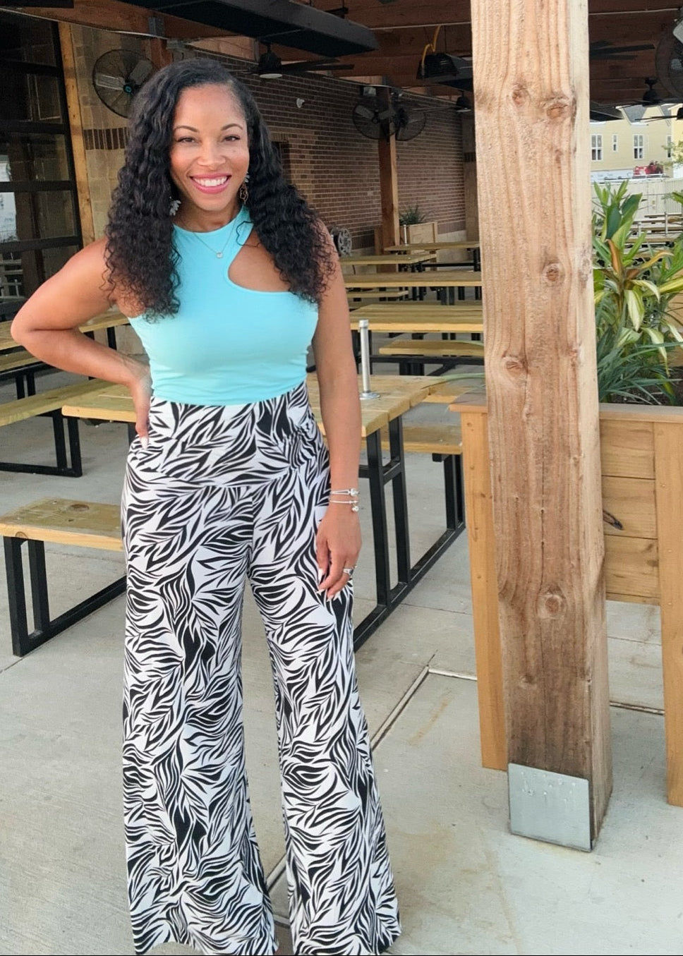 Woman in a blue top and patterned pants standing in an outdoor setting with wooden posts and tables.
