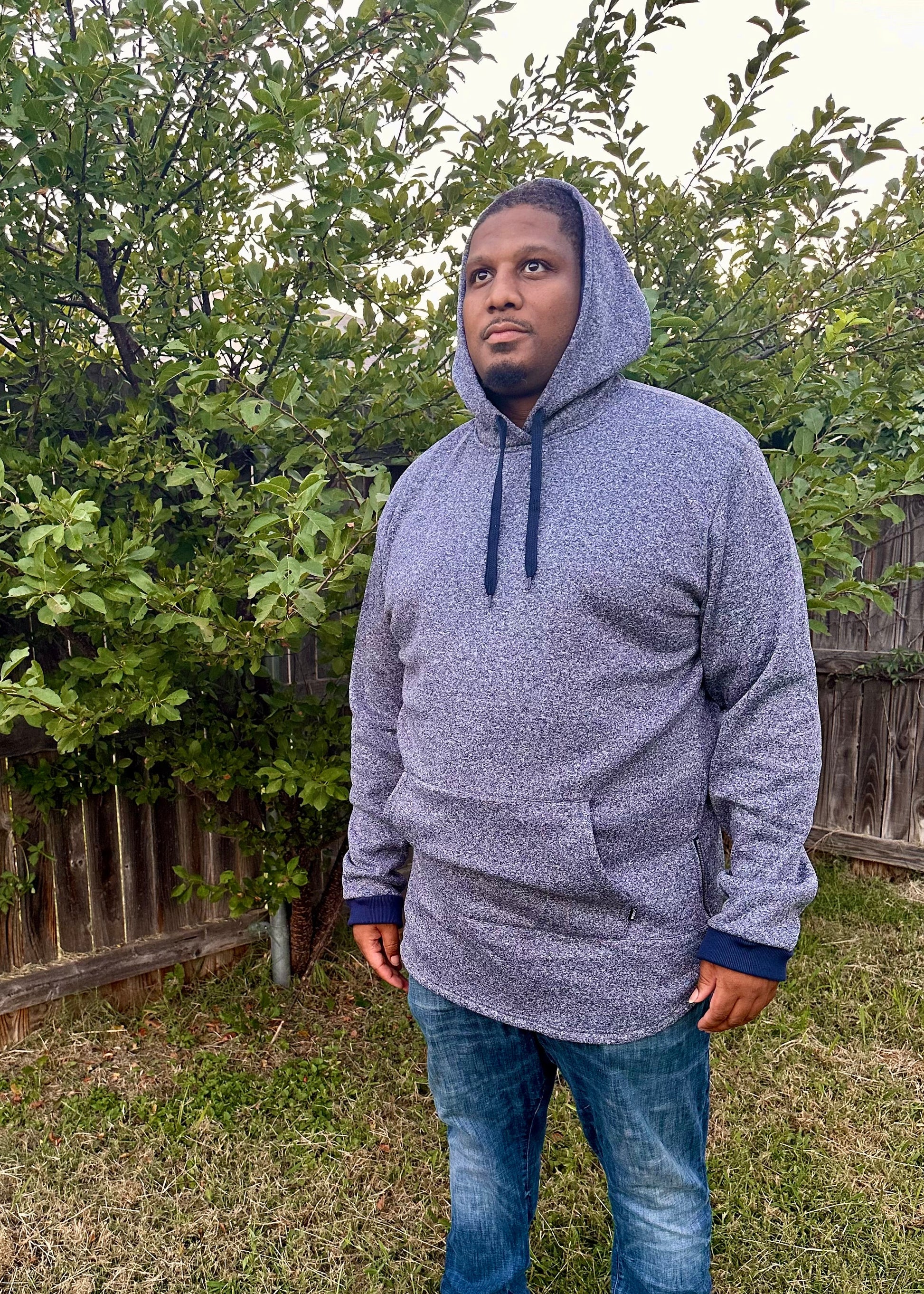Person wearing a gray hoodie and blue jeans standing in a backyard with greenery.