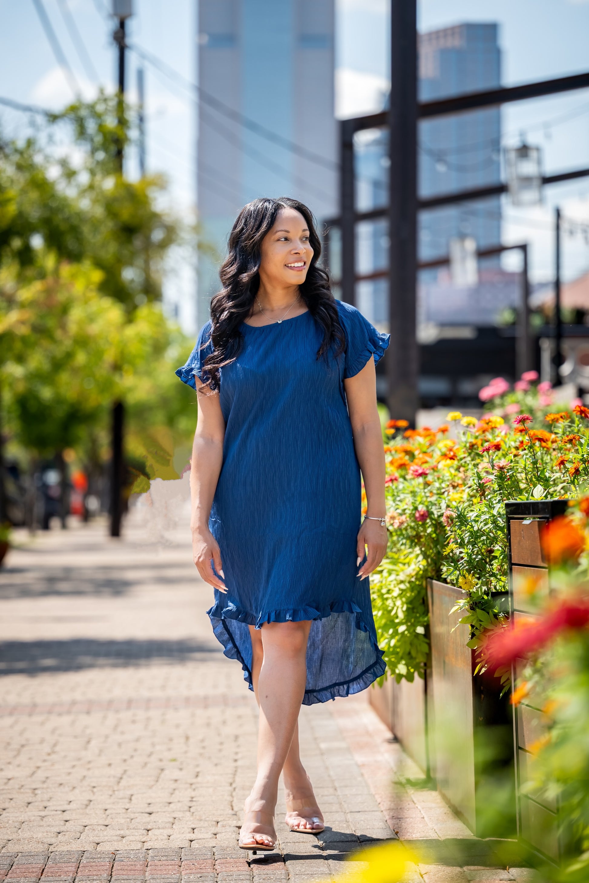 Woman in a blue dress walking on a sidewalk with flowers and buildings in the background