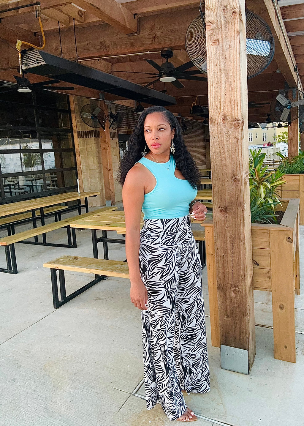Woman in a light blue top and black and white patterned skirt standing in an outdoor setting with wooden benches and plants.