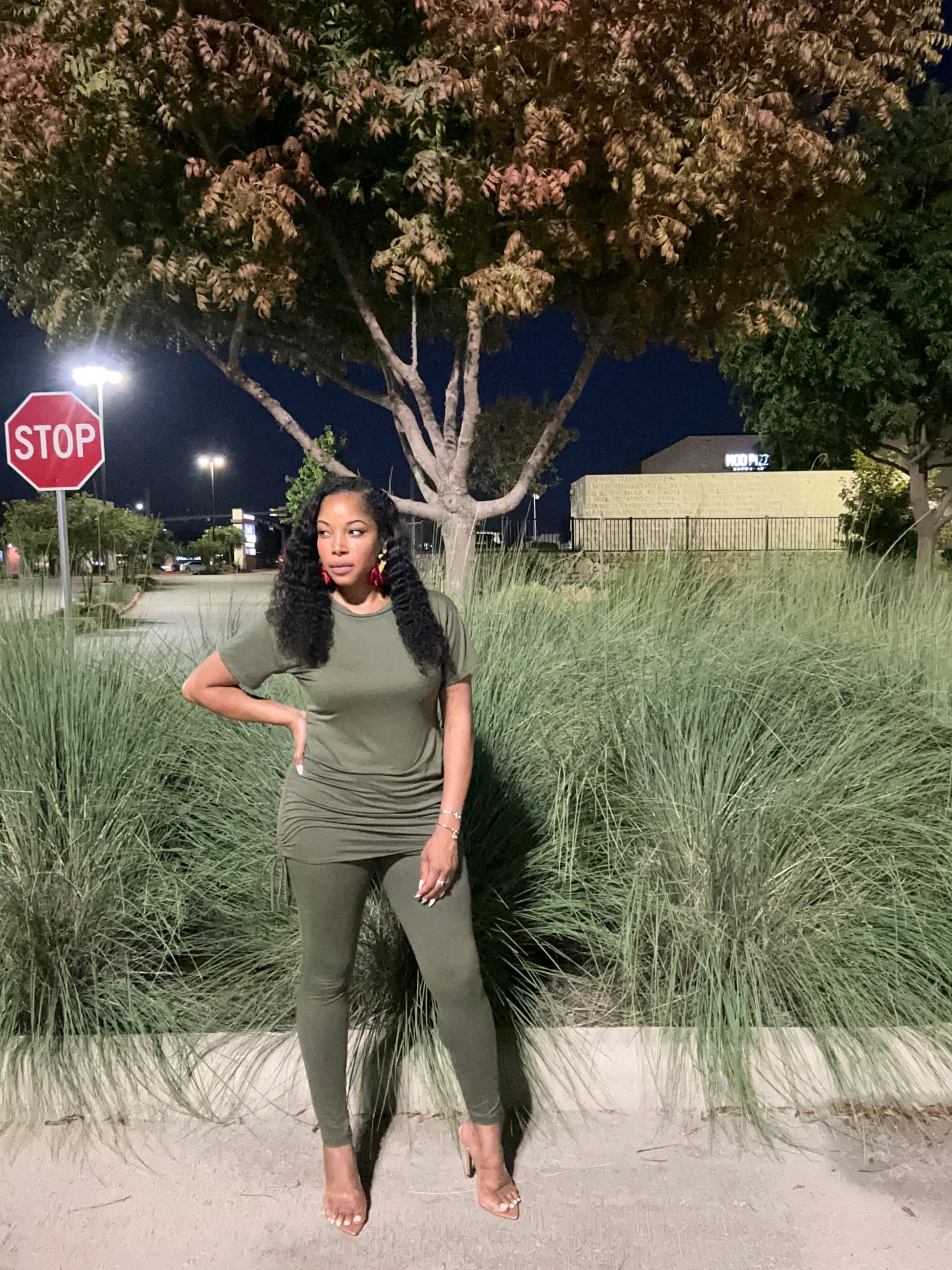 Woman in a green outfit standing outdoors at night with a tree and stop sign in the background.