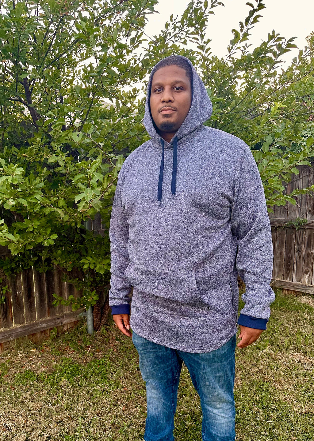 Person wearing a gray hoodie and blue jeans standing in a backyard with greenery.