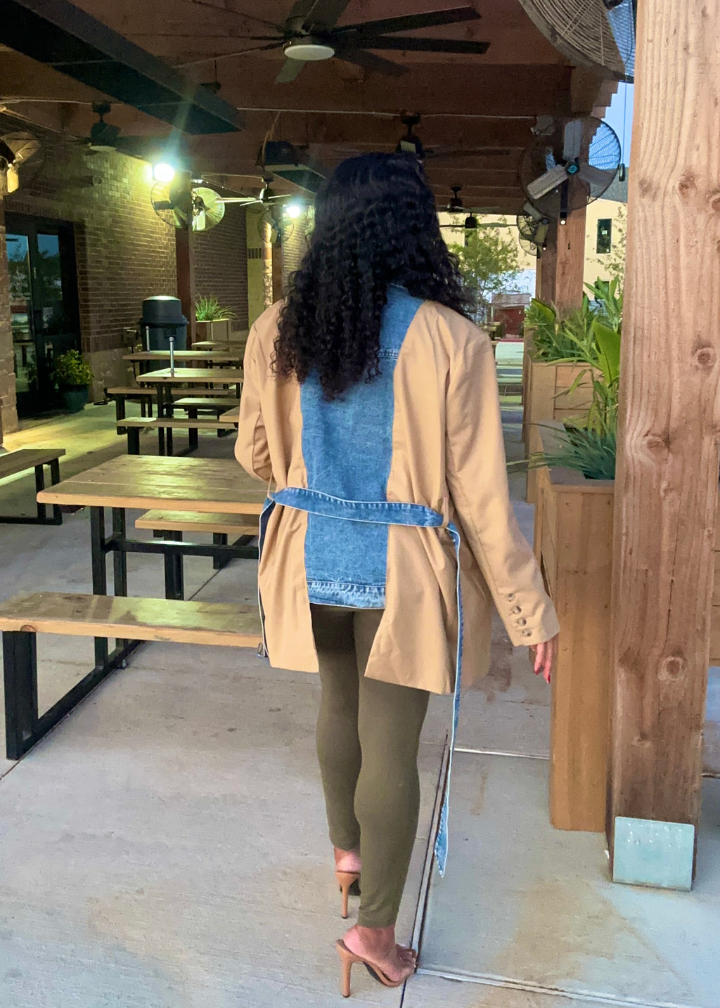 Person wearing a beige coat over a denim vest with green pants, standing on a patio.