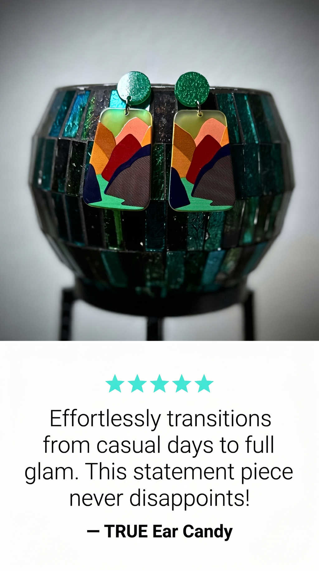 A pair of rectangular statement earrings with a colorful geometric mountain pattern in orange, green, brown, and black, hanging from green round studs. The earrings are displayed on a black stand in front of a dark green faceted glass bowl, with a promotional text block below.